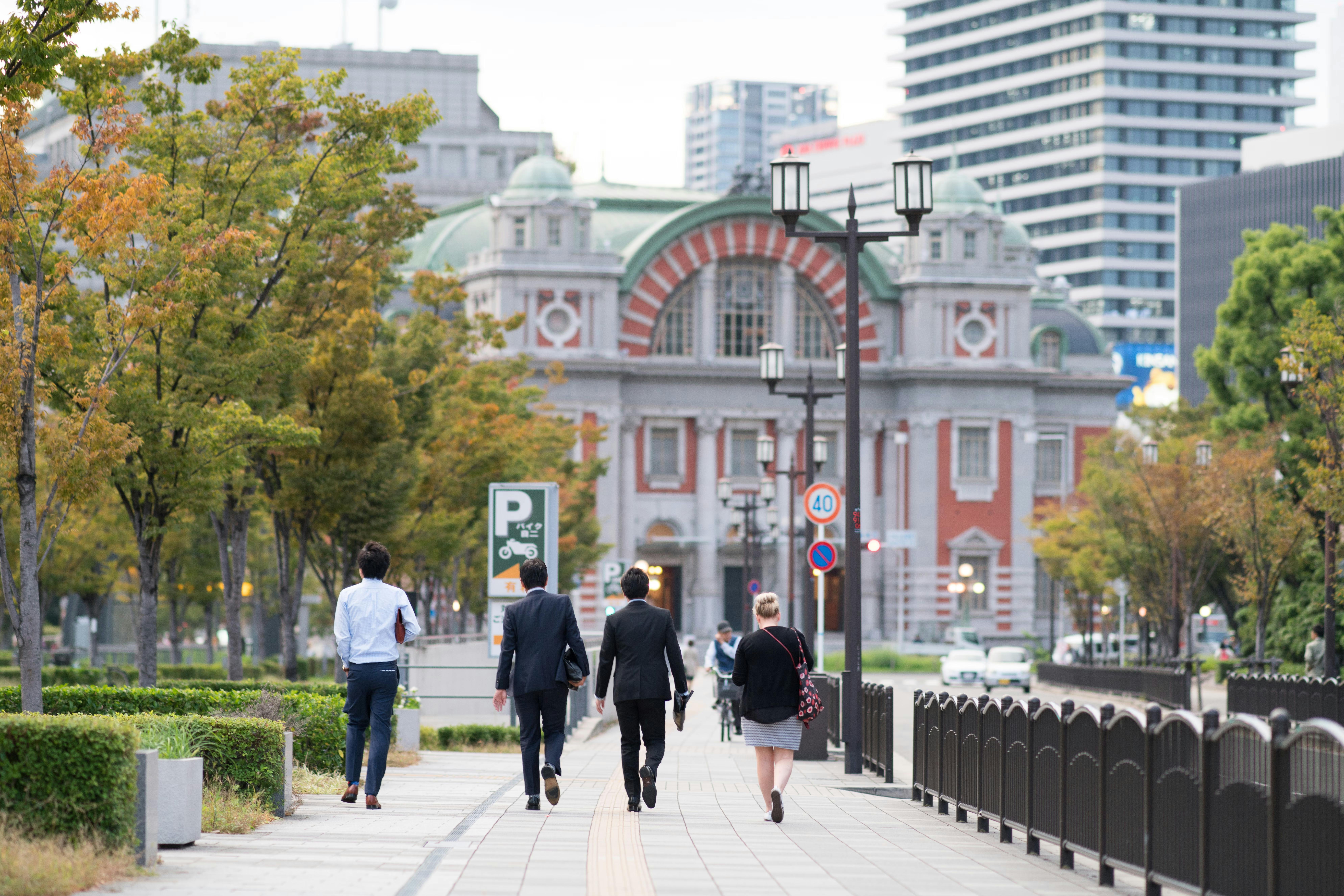 Four people walk down a tree-lined city sidewalk toward a historic red-brick building with a green roof. Modern high-rise buildings are visible in the background.