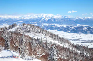 Snow-covered mountains and forested hills stretch across the landscape under a clear blue sky. The valley below is blanketed in white, with distant mountains rising majestically in the background.