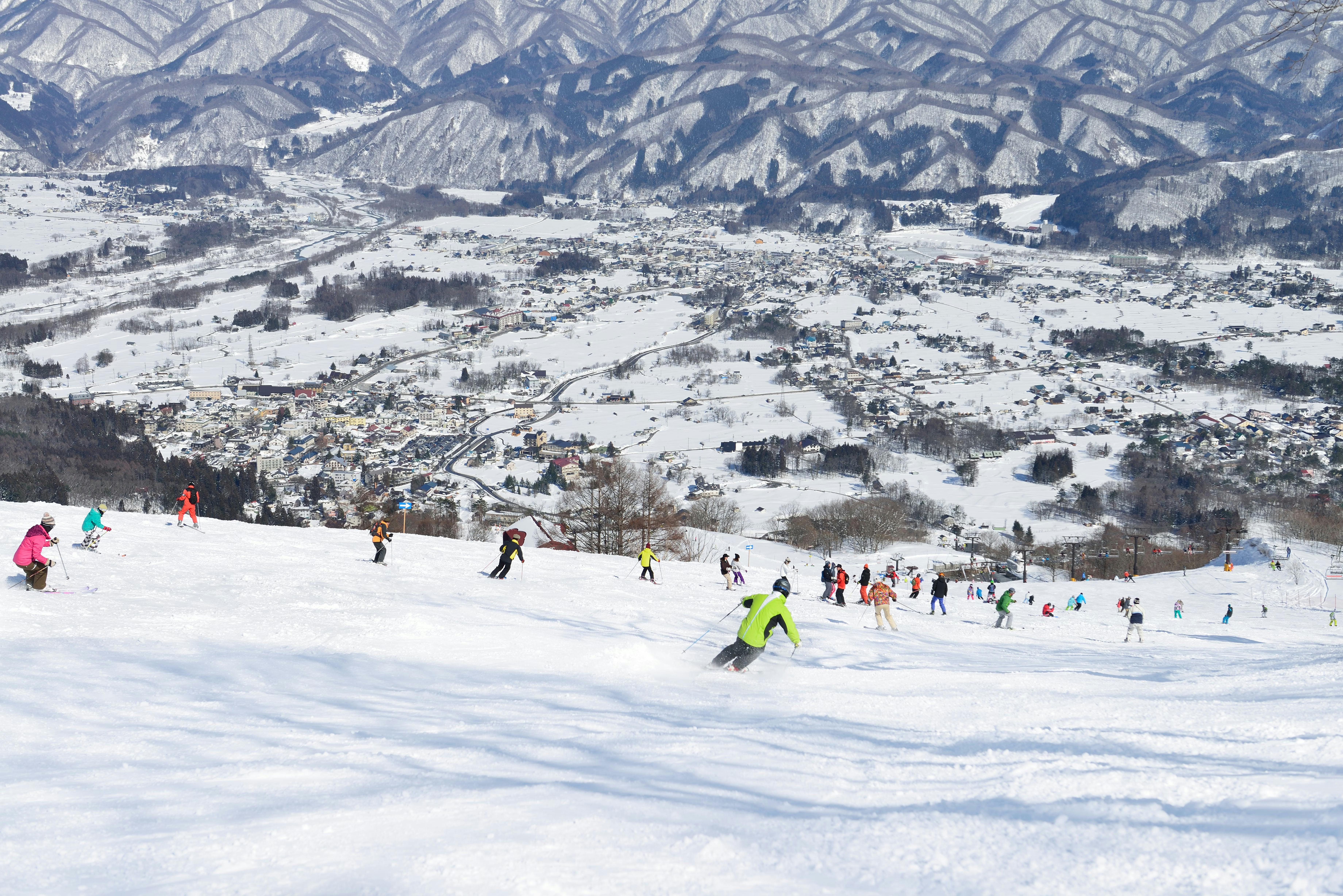 Skiers descend a snowy slope at a ski resort, with a snow-covered town and mountain range visible in the background under a clear sky.