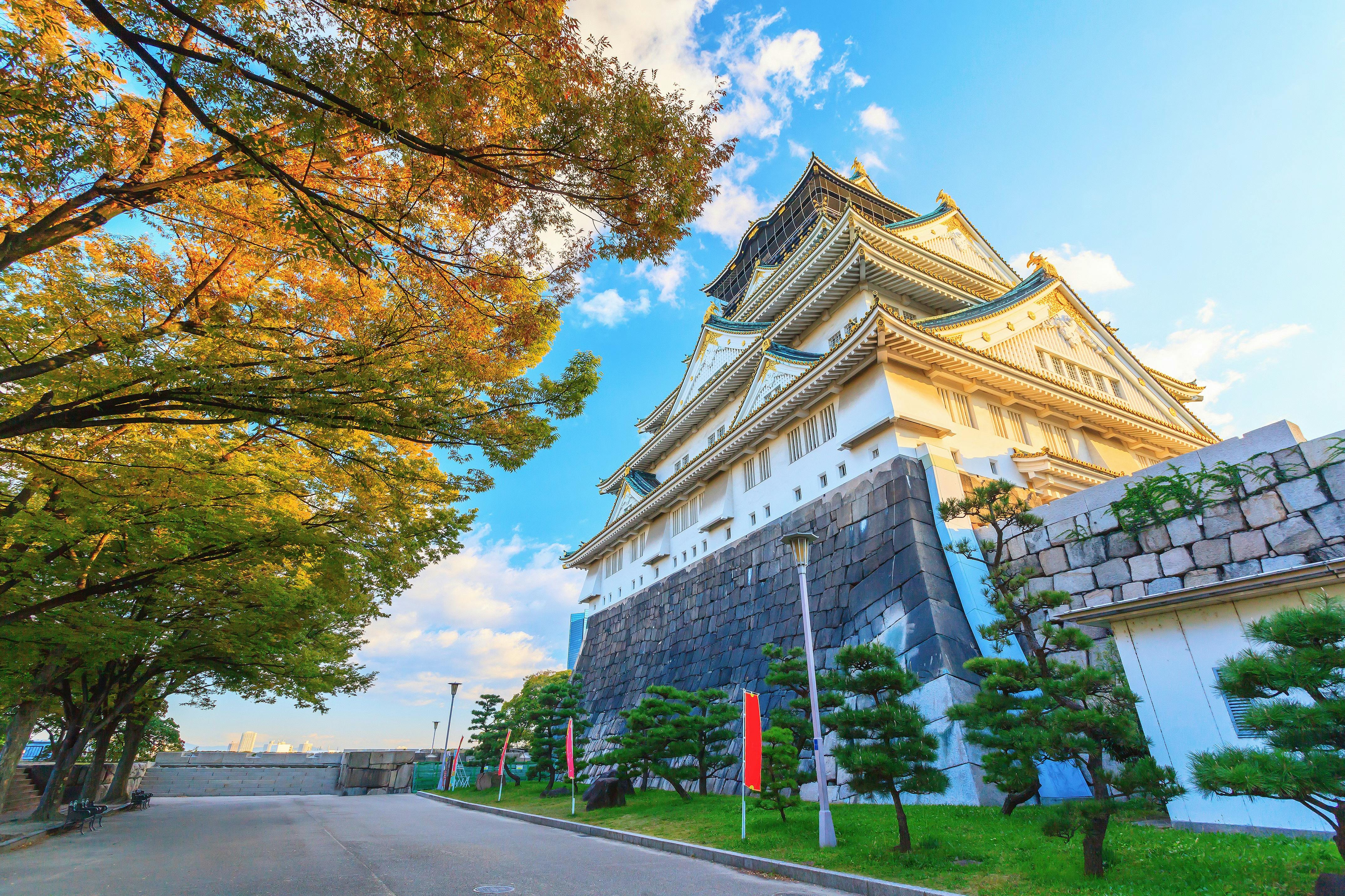 A traditional Japanese castle with white walls and dark tiled roofs stands against a blue sky, surrounded by green trees and a stone wall, with a path beside it.