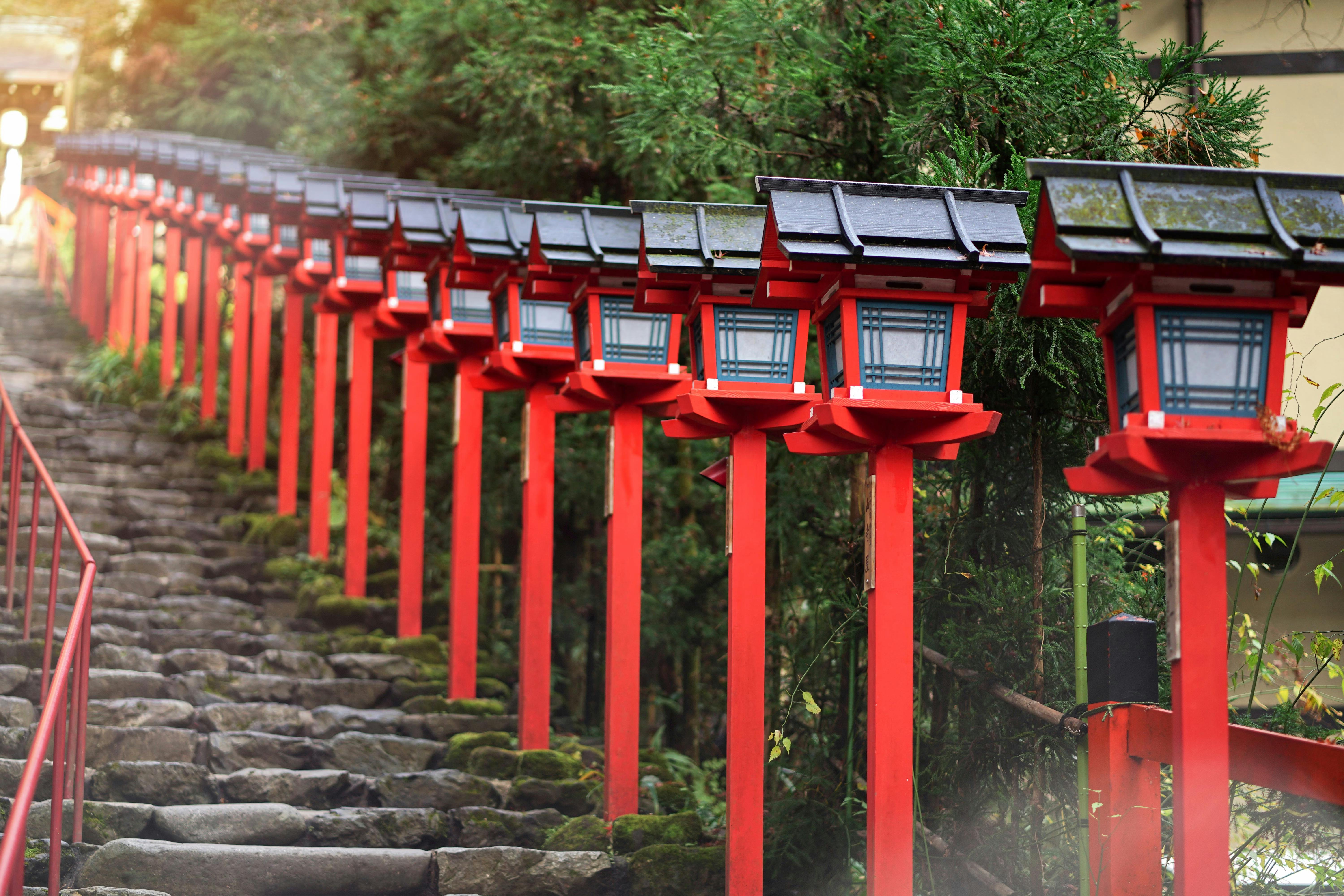 A row of traditional red Japanese lanterns lines a stone staircase surrounded by greenery, creating a serene and inviting pathway.