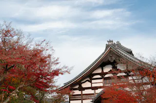 Traditional Japanese temple with ornate roof, surrounded by vibrant red autumn leaves and trees, set against a blue sky with wispy clouds.
