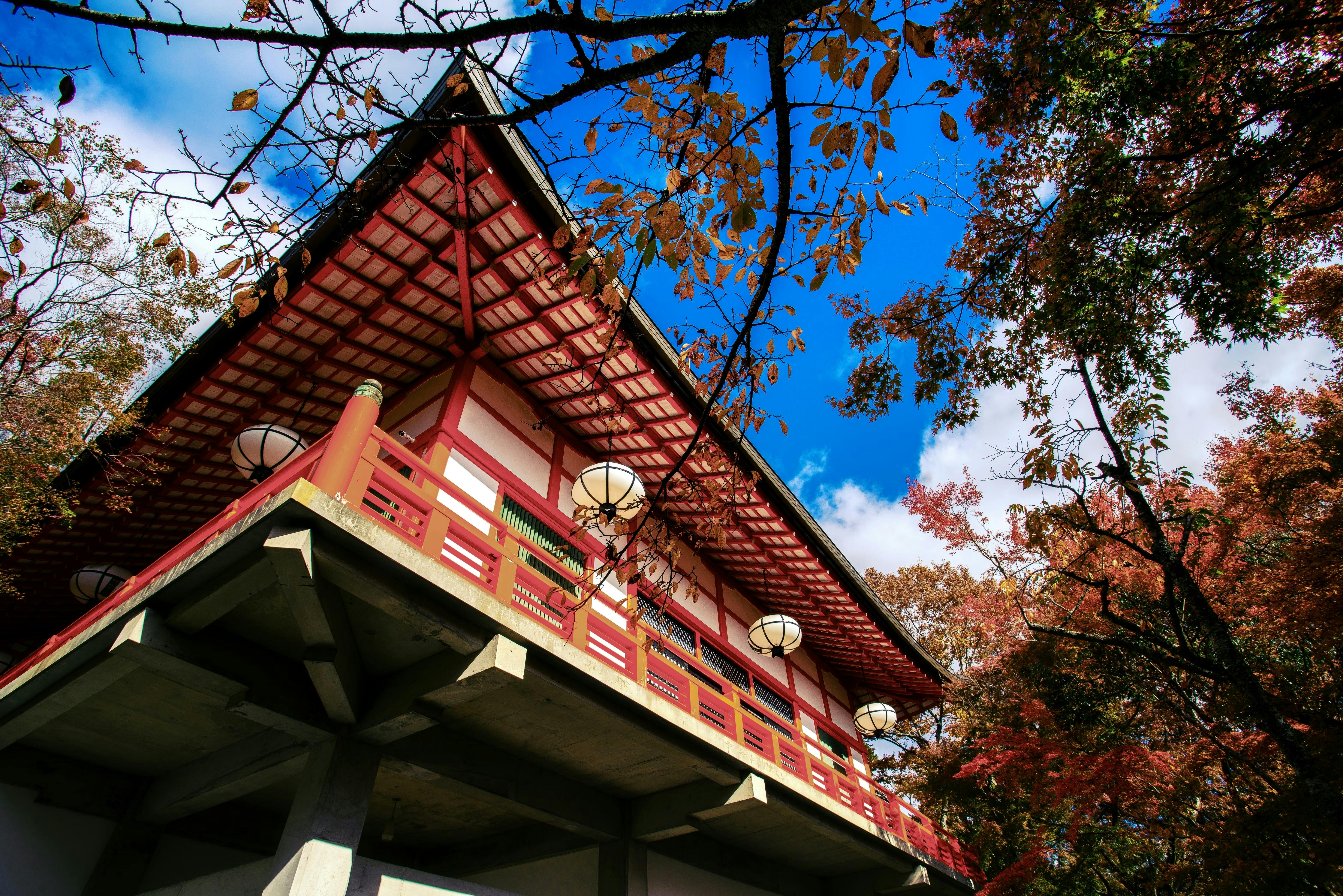 A traditional Japanese building with a red and white exterior is surrounded by trees with autumn leaves under a bright blue sky with scattered clouds.