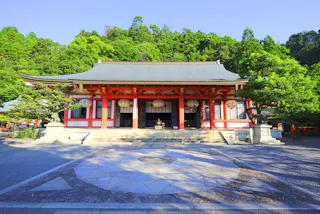 A traditional Japanese shrine with red pillars and a grey-tiled roof stands in front of lush green trees under a clear blue sky, with a patterned stone courtyard in the foreground.