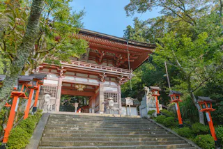 Stone steps lead up to a traditional Japanese shrine gate surrounded by green trees, red lanterns, and guardian statues on each side, with sunlight illuminating the structure.