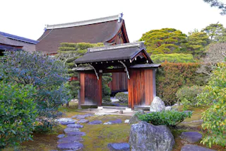 A traditional Japanese wooden gate stands in a lush garden with stone paths, manicured bushes, and a wooden building with a tiled roof in the background.