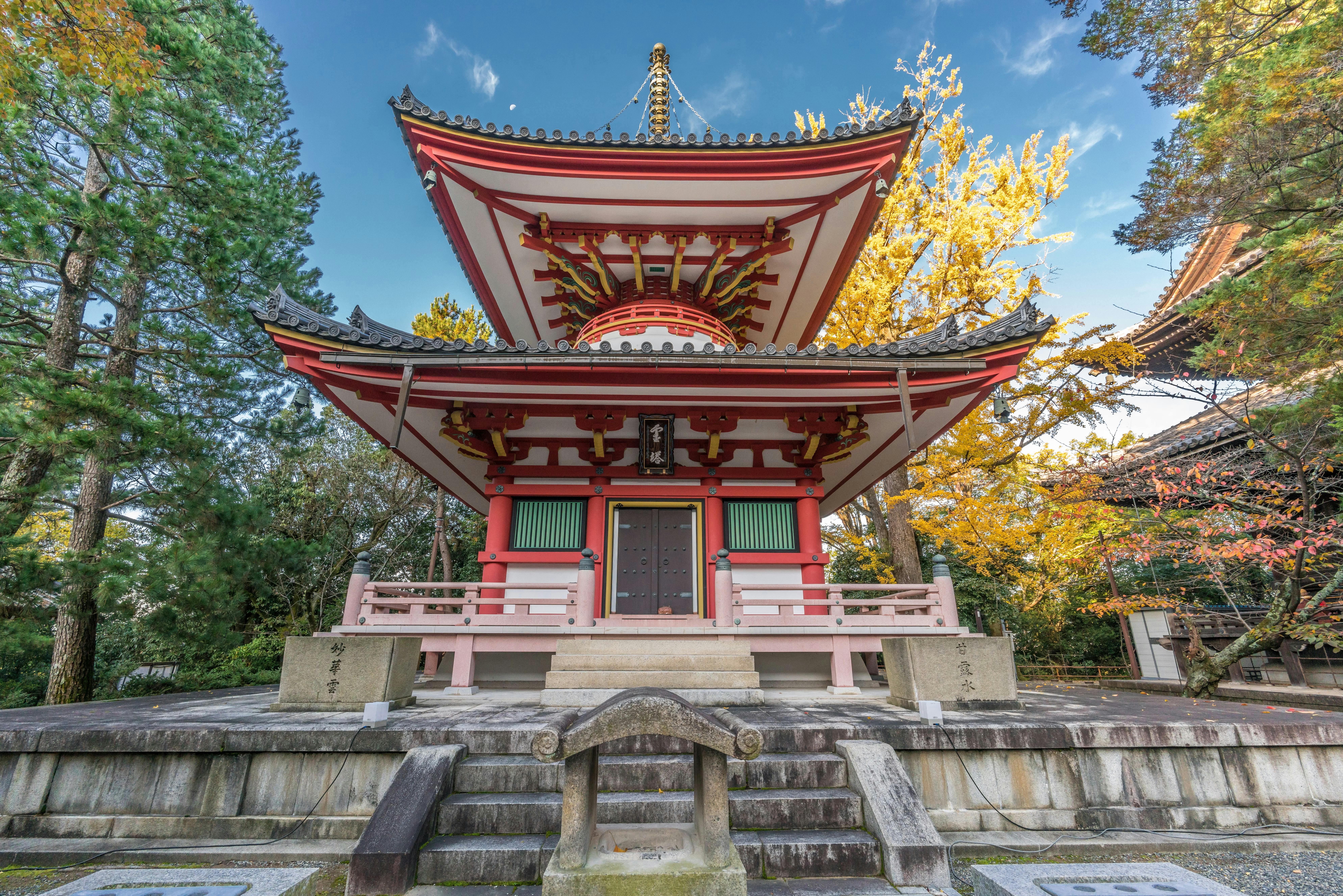 A traditional Japanese pagoda with red and white detailing stands among green and yellow trees on a clear day, with a stone path and small archway in front.
