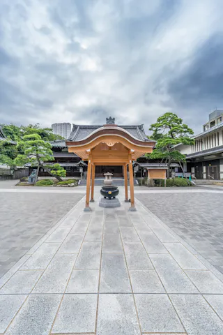 A traditional Japanese structure with a wooden roof stands in the center of a paved courtyard, surrounded by manicured trees and temple buildings under a cloudy sky.