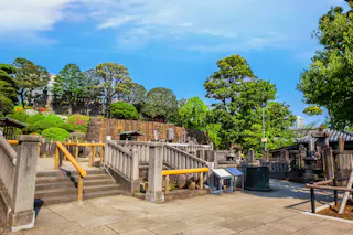 Stone steps and pathways surrounded by lush green trees and shrubs in a traditional Japanese garden under a bright blue sky.