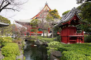 A traditional Japanese garden with a small pond, manicured bushes, and a red wooden pavilion, set in front of a large temple building with a sloped roof and ornate details.