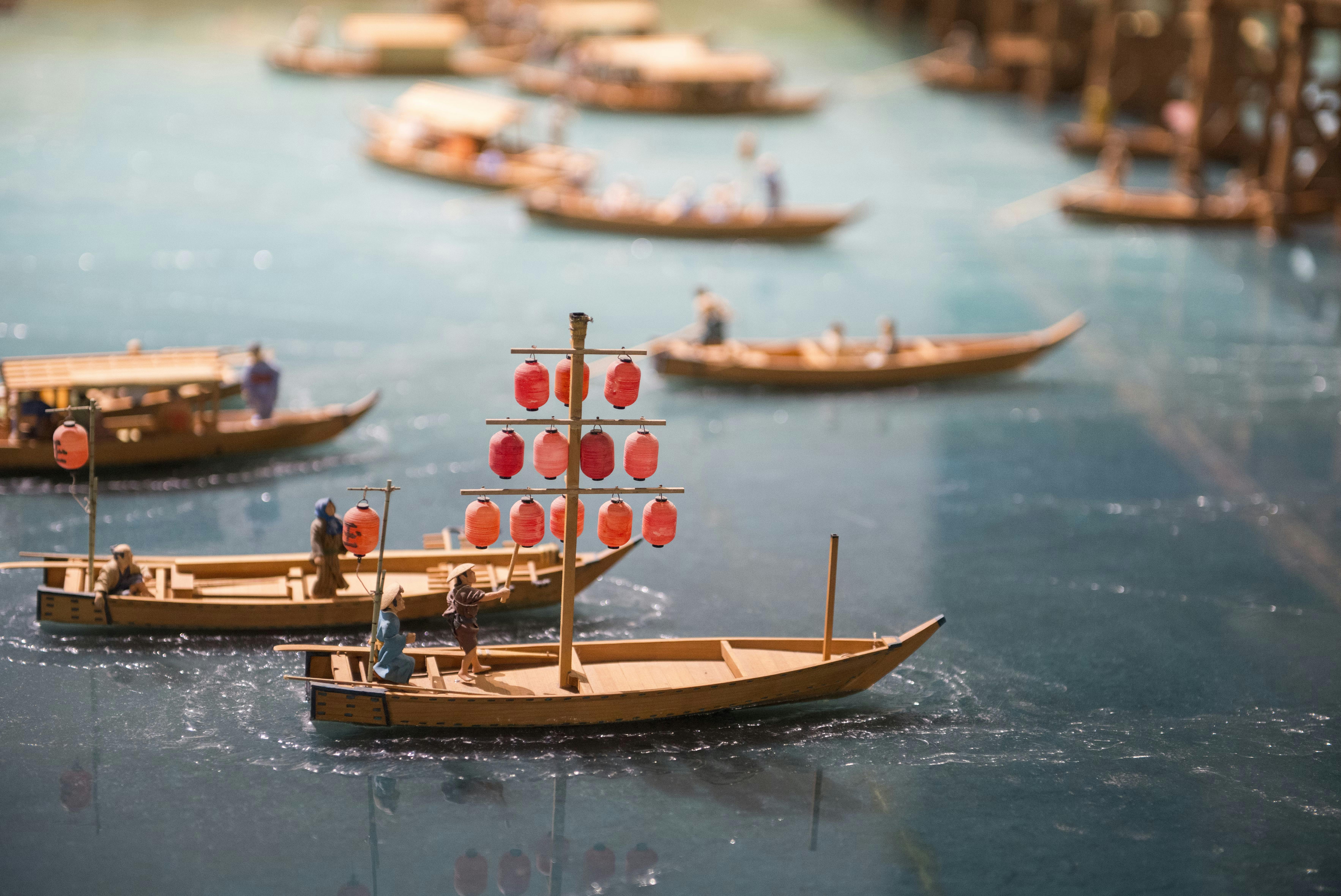Model boats with miniature figures float on a blue water surface. One boat in the foreground displays red lanterns. Other boats and figures are visible in the background, creating a lively scene.