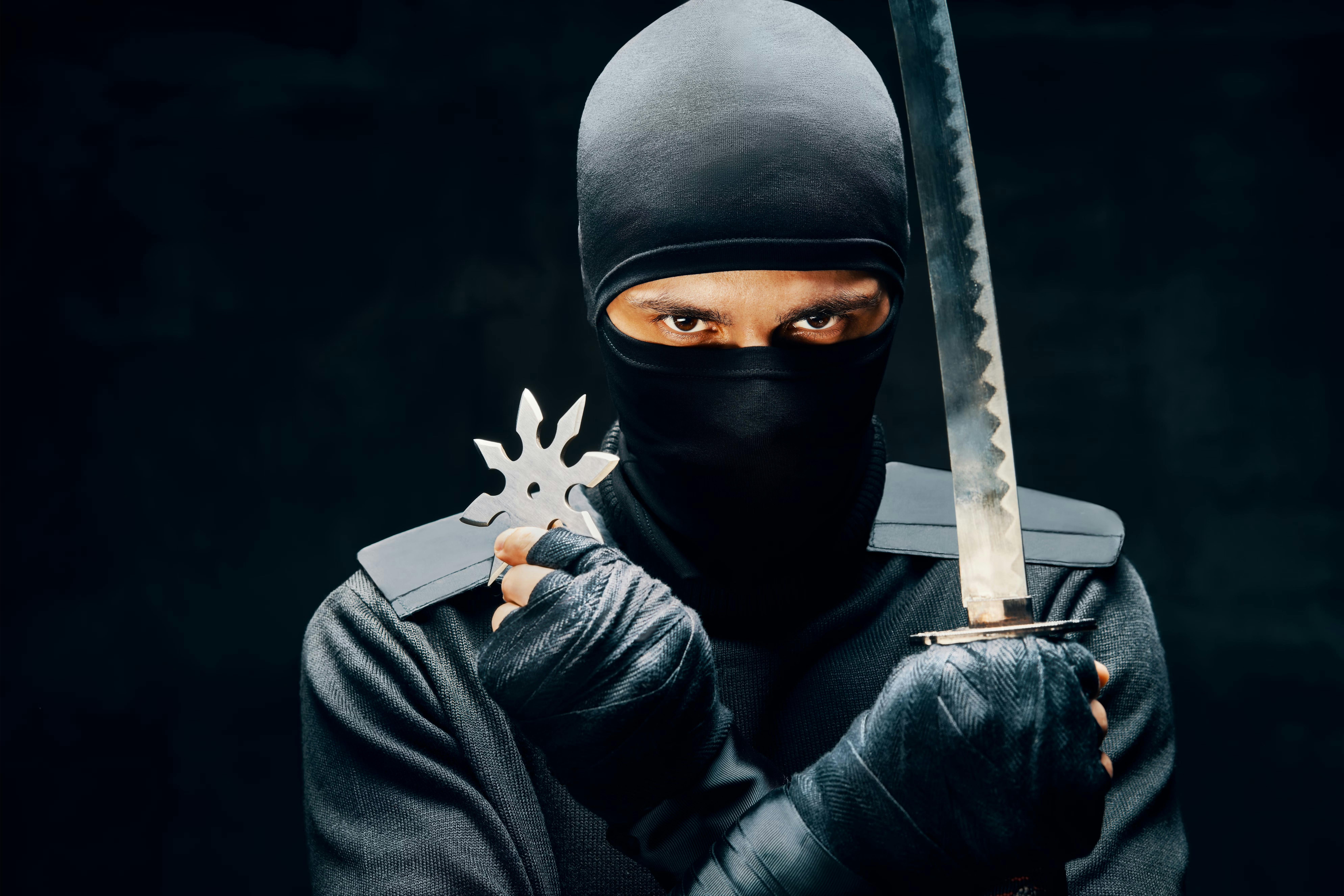 A person dressed in black ninja attire, holding a katana sword in one hand and a shuriken in the other, with only their intense eyes visible through the mask against a dark background.