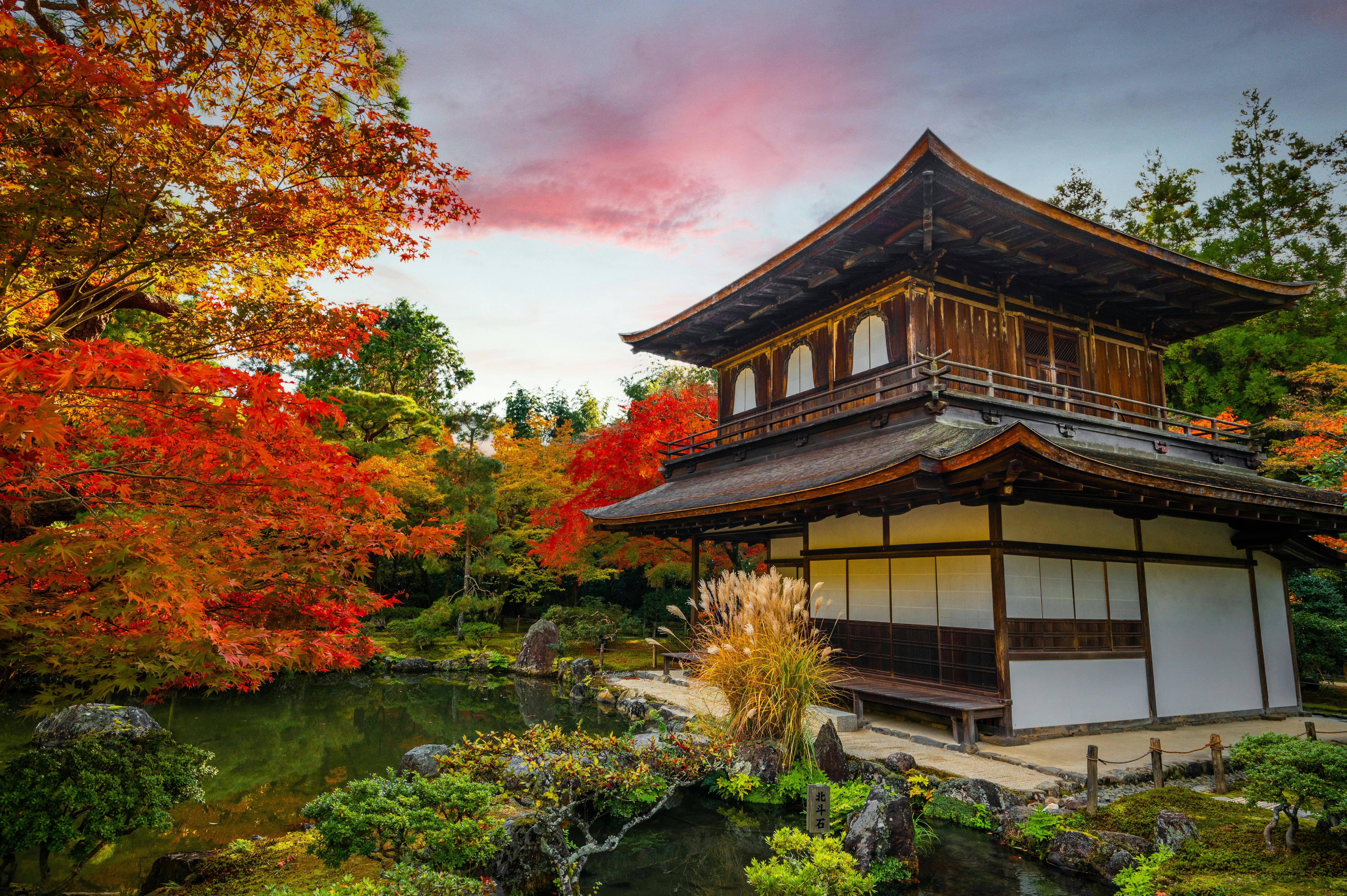 A traditional Japanese wooden building stands beside a tranquil pond, surrounded by vibrant red and orange autumn foliage, under a colorful sky at dusk.