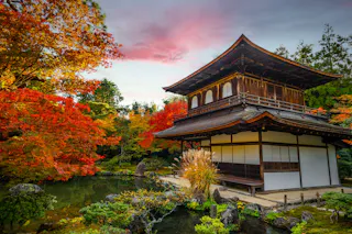 A traditional Japanese wooden building stands beside a tranquil pond, surrounded by vibrant red and orange autumn foliage, under a colorful sky at dusk.