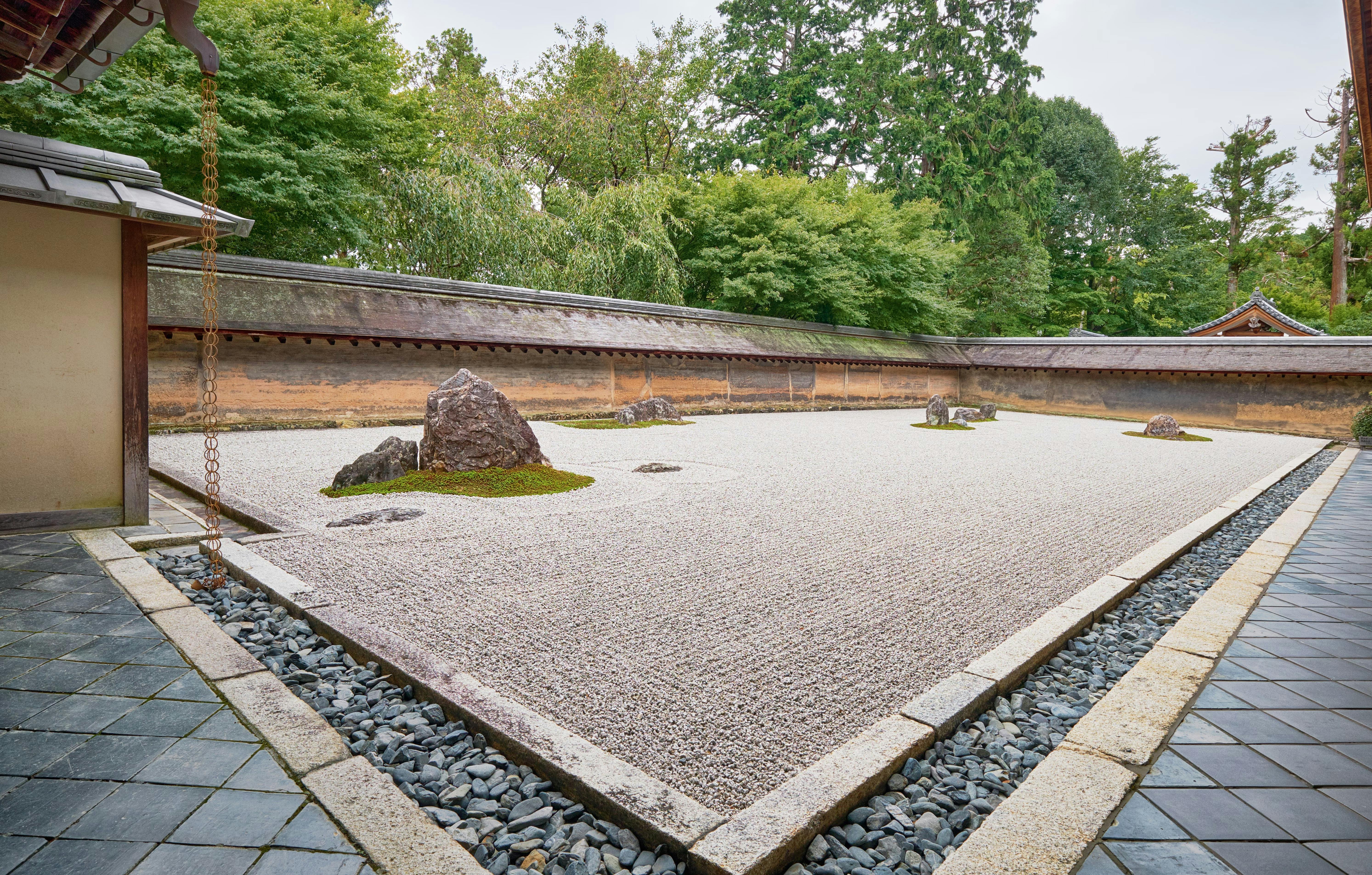 A traditional Japanese rock garden with raked white gravel, scattered large rocks, and patches of moss, surrounded by a stone border and greenery with a wooden wall in the background.