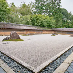 Ryoan-ji Temple A traditional Japanese rock garden with raked white gravel, scattered large rocks, and patches of moss, surrounded by a stone border and greenery with a wooden wall in the background.