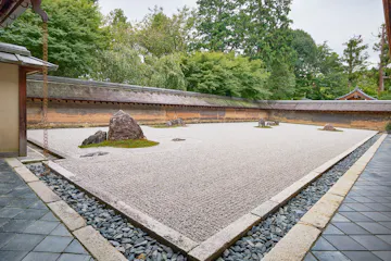 Ryoan-ji Temple A traditional Japanese rock garden with raked white gravel, scattered large rocks, and patches of moss, surrounded by a stone border and greenery with a wooden wall in the background.
