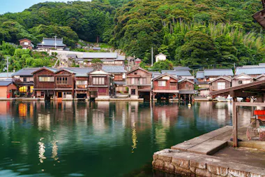 Traditional wooden houses line the waterfront, built on stilts over calm water, with lush green forested hills rising in the background under a cloudy sky. Some boats are docked near the houses.