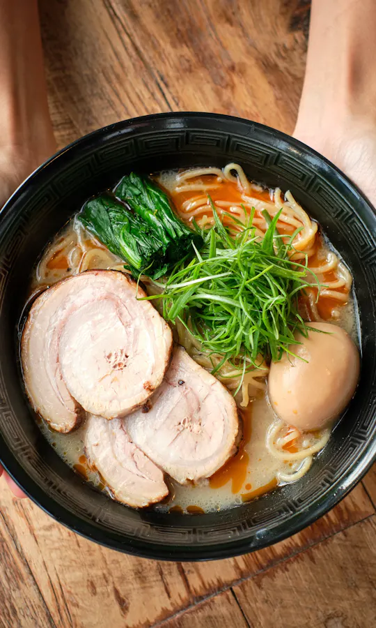 Ramen A person holds a black bowl filled with ramen topped with sliced pork, a boiled egg, green onions, spinach, and noodles, above a wooden table.