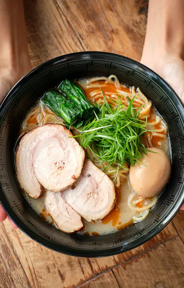 Ramen A person holds a black bowl filled with ramen topped with sliced pork, a boiled egg, green onions, spinach, and noodles, above a wooden table.