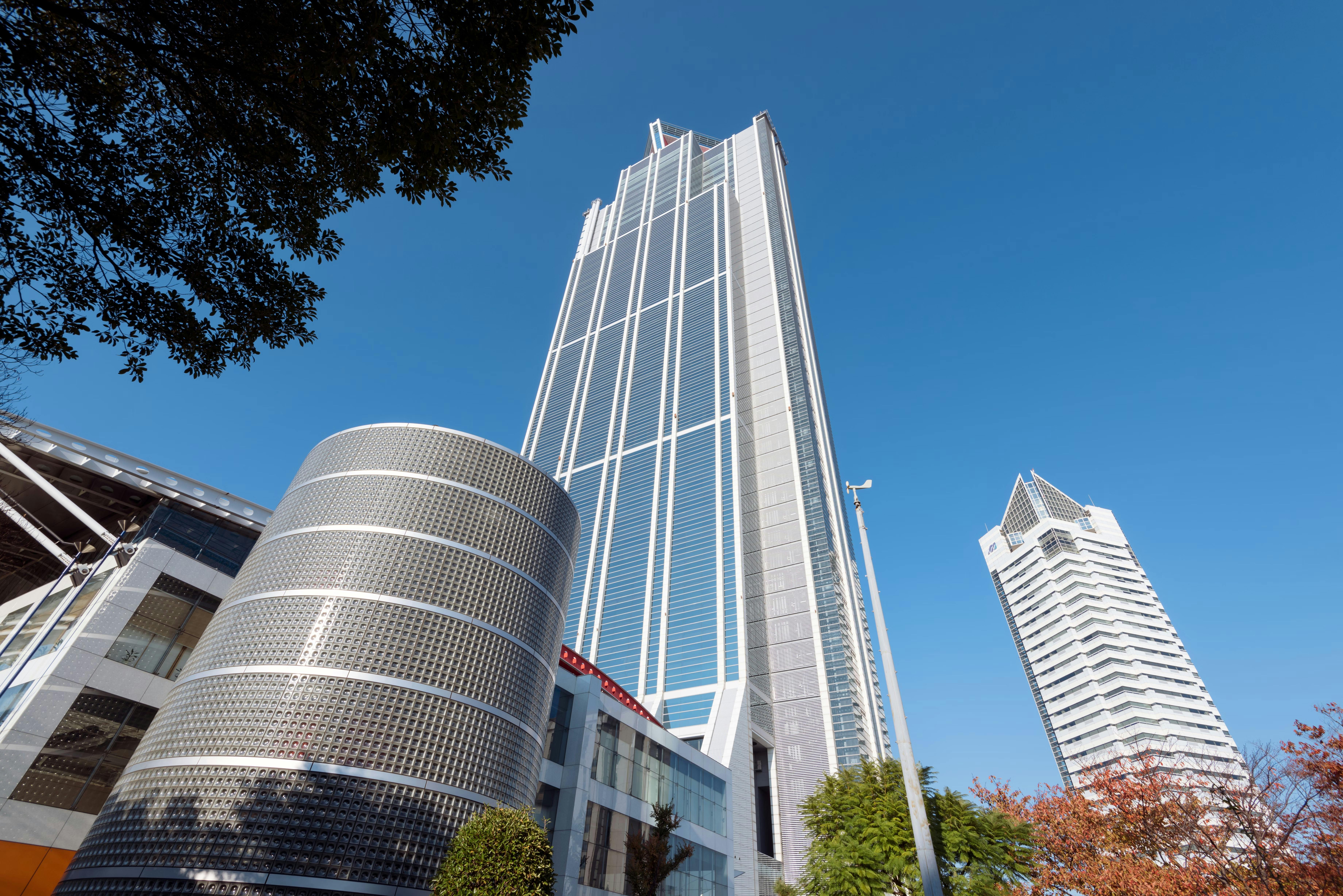 Tall modern skyscraper with a glass facade rises against a clear blue sky, flanked by a cylindrical building with a patterned exterior and another high-rise with a unique white rooftop structure. Trees border the foreground.