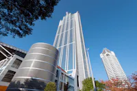Tall modern skyscraper with a glass facade rises against a clear blue sky, flanked by a cylindrical building with a patterned exterior and another high-rise with a unique white rooftop structure. Trees border the foreground.