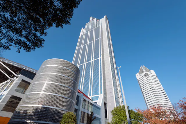 Tall modern skyscraper with a glass facade rises against a clear blue sky, flanked by a cylindrical building with a patterned exterior and another high-rise with a unique white rooftop structure. Trees border the foreground.