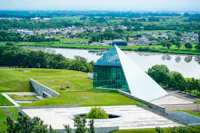 A modern glass pyramid building sits on a grassy landscape near a river, with trees and a distant town visible in the background under a hazy sky.