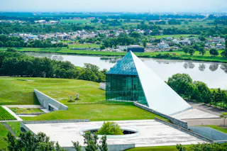 A modern glass pyramid building sits on a grassy landscape near a river, with trees and a distant town visible in the background under a hazy sky.
