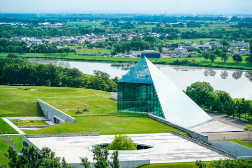 A modern glass pyramid building sits on a grassy landscape near a river, with trees and a distant town visible in the background under a hazy sky.