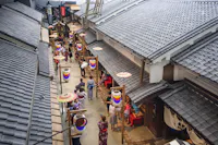 A view from above shows people walking along a narrow street lined with traditional Japanese buildings and colorful lanterns, some visitors wearing yukatas and kimonos.