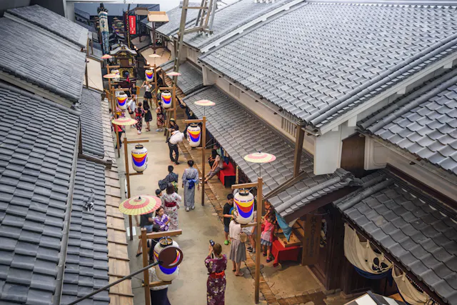A view from above shows people walking along a narrow street lined with traditional Japanese buildings and colorful lanterns, some visitors wearing yukatas and kimonos.