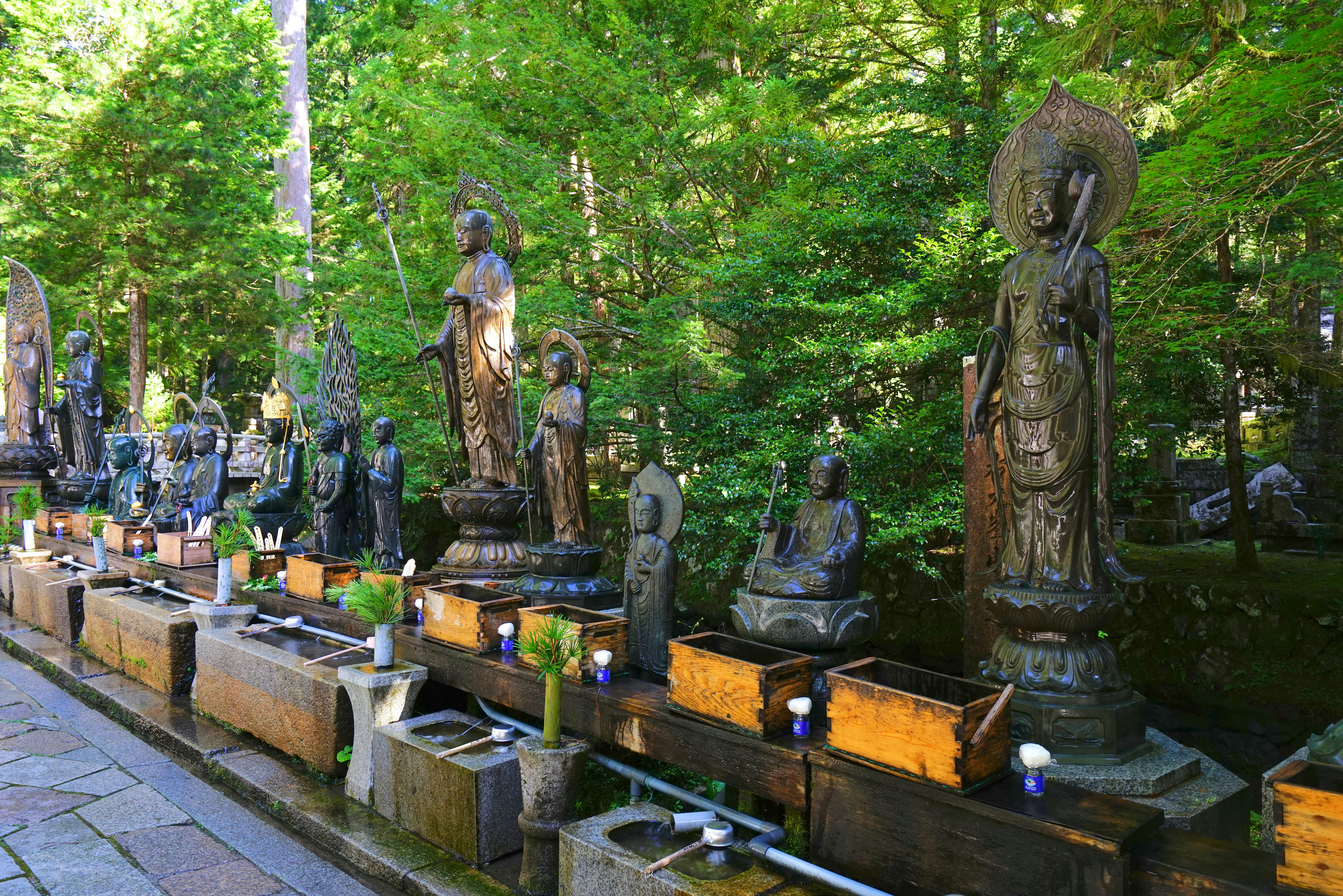 A row of bronze Buddhist statues of various sizes stands on stone platforms surrounded by lush green trees, with small wooden boxes and ritual objects placed in front of each statue.