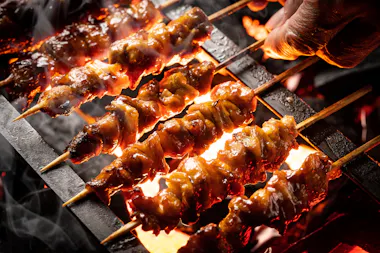 Close-up of skewered meat being grilled over an open flame, with a hand turning one of the skewers. The meat is glazed and sizzling, and smoke rises from the hot grill.