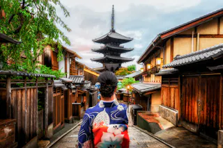 A person wearing a colorful kimono walks down a traditional street lined with wooden buildings, facing a tall pagoda in the distance under a partly cloudy sky.