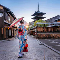 Gion A woman in a colorful kimono holding a parasol walks down a quiet stone street in Kyoto, Japan, with traditional wooden buildings and a pagoda visible in the background at sunset.