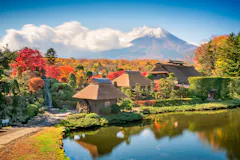Traditional Japanese thatched-roof houses by a pond, surrounded by vibrant autumn foliage, with Mt. Fuji in the background under a blue sky with white clouds.