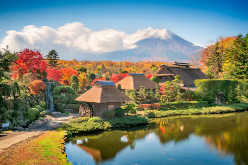 Traditional Japanese thatched-roof houses by a pond, surrounded by vibrant autumn foliage, with Mt. Fuji in the background under a blue sky with white clouds.