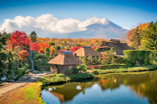 Traditional Japanese thatched-roof houses by a pond, surrounded by vibrant autumn foliage, with Mt. Fuji in the background under a blue sky with white clouds.