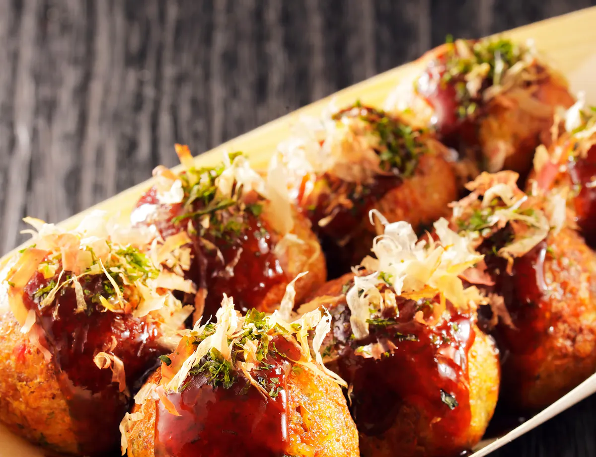 Takoyaki A wooden tray holds several pieces of takoyaki, Japanese octopus balls, topped with brown sauce, green seaweed, and bonito flakes, set on a dark wooden surface.