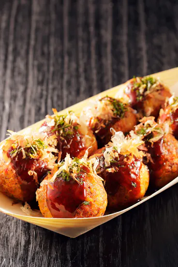 A wooden tray holds several pieces of takoyaki, Japanese octopus balls, topped with brown sauce, green seaweed, and bonito flakes, set on a dark wooden surface.