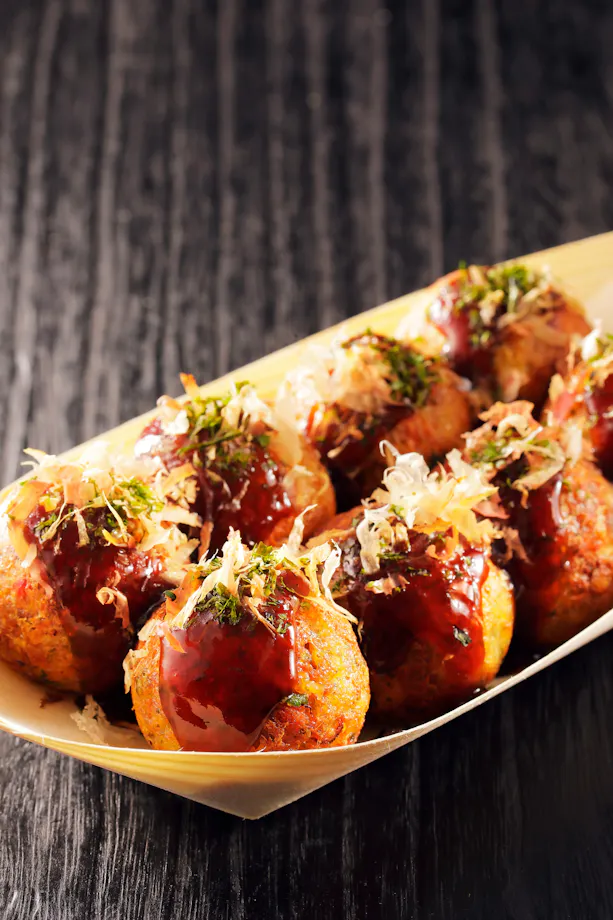 A wooden tray holds several pieces of takoyaki, Japanese octopus balls, topped with brown sauce, green seaweed, and bonito flakes, set on a dark wooden surface.
