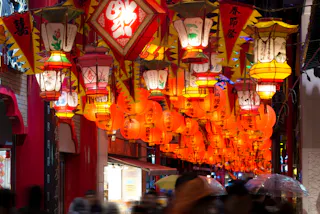 A vibrant street scene filled with colorful Chinese lanterns and illuminated signs, with people walking beneath, some holding umbrellas. The lanterns feature various Chinese characters and traditional designs.