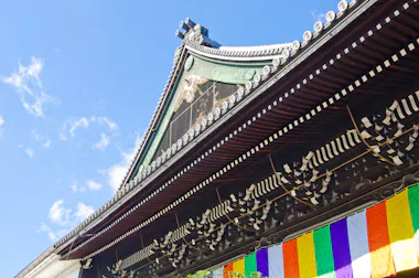Close-up of a traditional Japanese temple roof with ornate wooden carvings, under a blue sky. Colorful vertical banners in red, white, green, yellow, and purple hang below the roof.