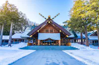 A traditional Japanese Shinto shrine with a wooden roof, large white banners featuring chrysanthemum crests, surrounded by snow and tall evergreen trees under a clear blue sky.