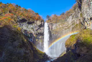 A tall waterfall cascades down a rocky cliff surrounded by autumn trees, with a bright rainbow arching through the mist at the base under a clear blue sky.