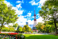 A tall red and white broadcast tower rises above lush green trees and colorful flower beds in a sunny park under a vibrant blue sky with scattered clouds.