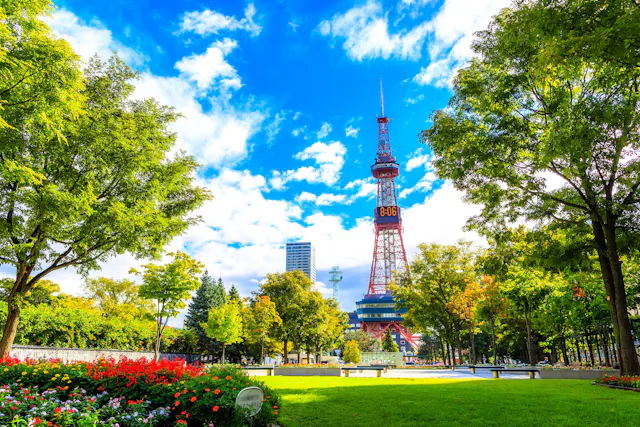 A tall red and white broadcast tower rises above lush green trees and colorful flower beds in a sunny park under a vibrant blue sky with scattered clouds.