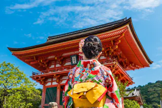 A woman in a colorful kimono with a yellow obi stands facing a traditional Japanese temple gate with red pillars and intricate roof details, under a bright blue sky.
