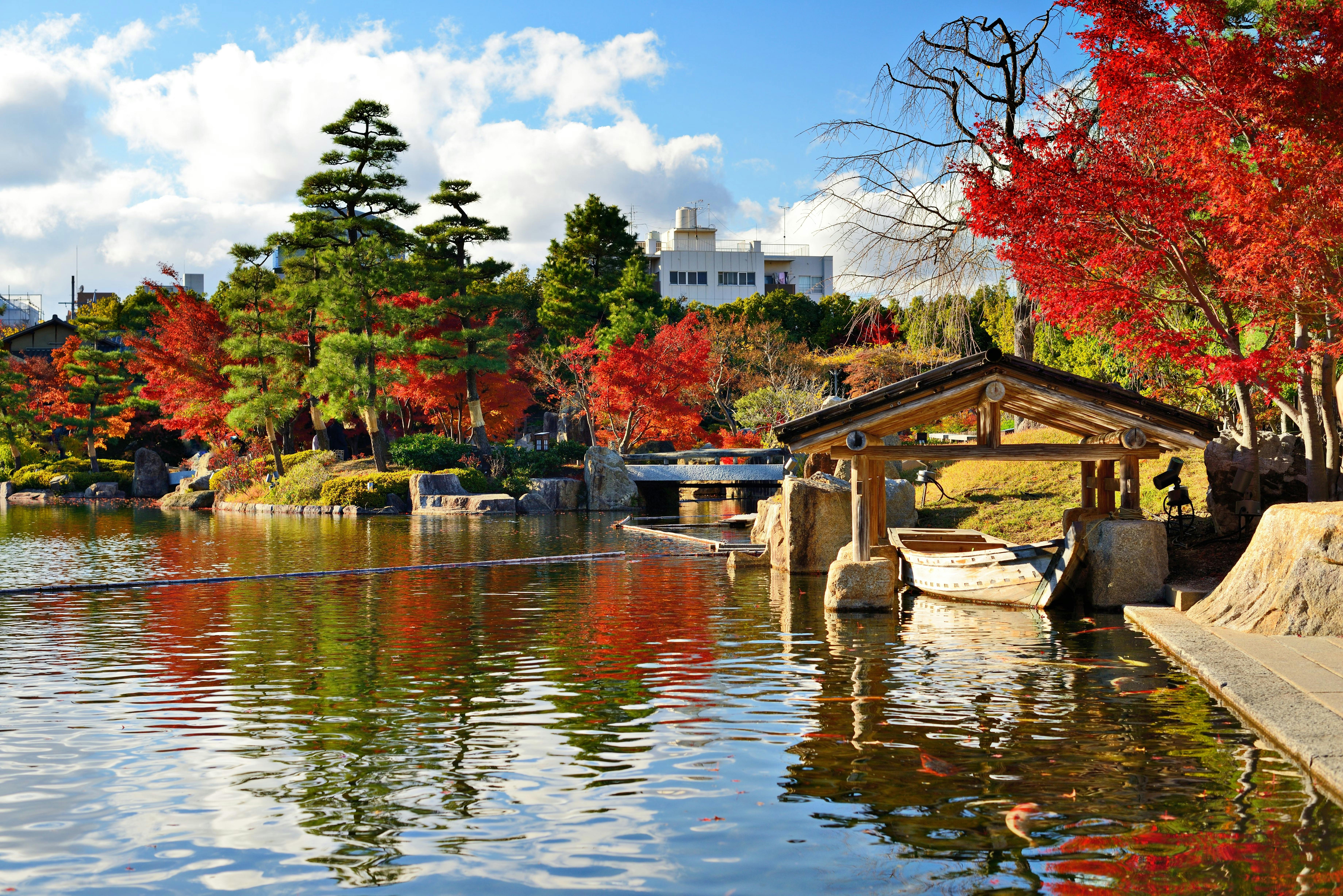 A serene Japanese garden with vibrant red and green trees reflected in a calm pond, a wooden boat dock, and a small shelter on the water’s edge under a partly cloudy sky.